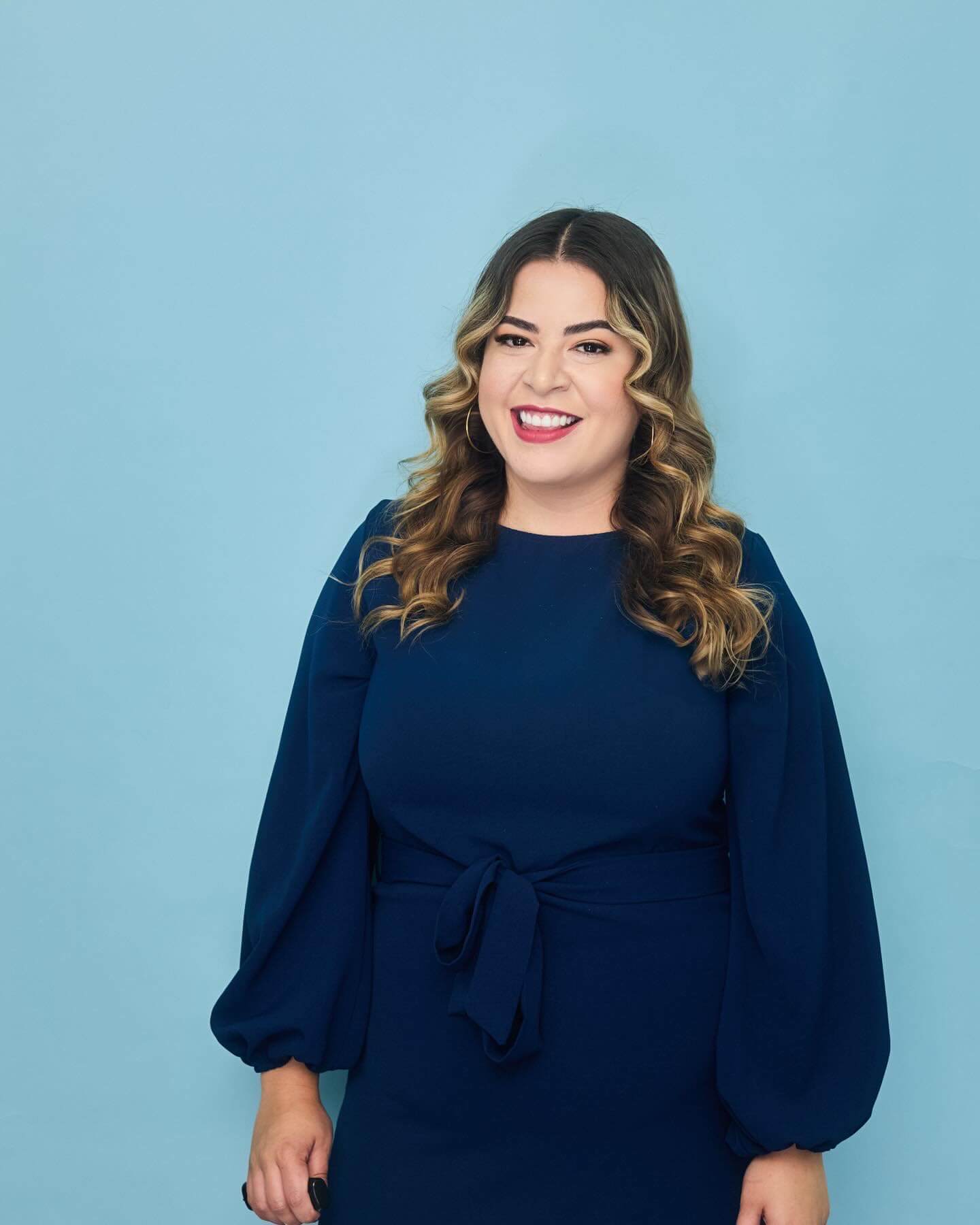 Photo featuring podcast audit client, Maria Rio. Maria smiling in front of a light blue background, wearing a navy blue dress with puffed sleeves and a tie at the waist. Her hair is styled in loose curls and she has hoop earrings.