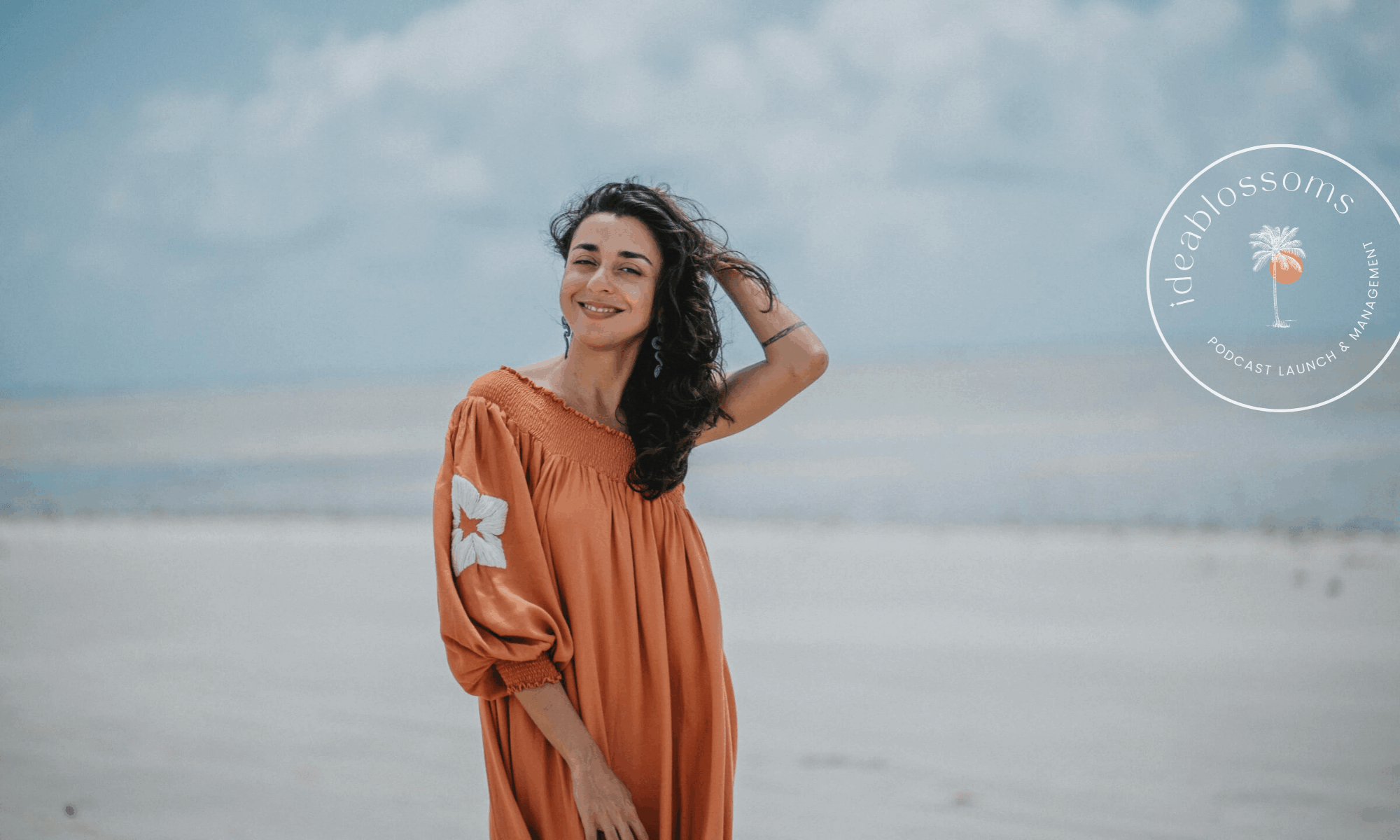 Rosa Sarmento, founder of Ideablossoms, standing on a misty beach wearing a loose rust-colored dress with white embroidered detail on the sleeve. She smiles softly while lifting her hair with one hand. The background features a muted blue-gray sky and shoreline. On the right side, a circular logo reads “ideablossoms – Podcast Launch & Management” with a small palm tree icon.